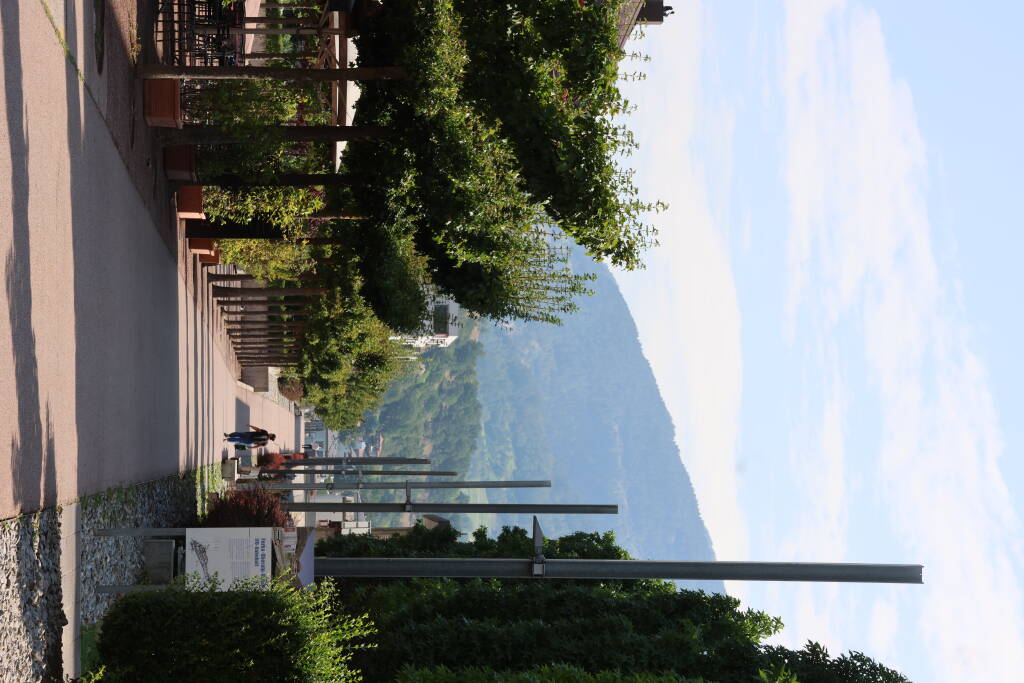 In place of the former railway is now pedestrian and cycling route through the Naters town. On the left, there is the former local station, now a café. Catenary poles are now serving for street lighting. A steep slope to the bridge is visible in the background.